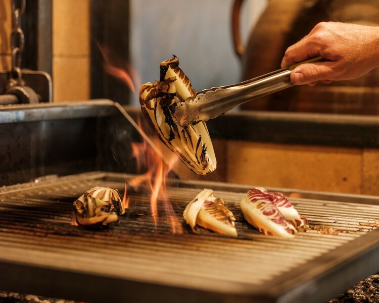 Grilled radicchio tardivo being turned over flames on a wood-fired grill, with tongs holding a charred section, showcasing the vegetable's distinctive red and white leaves.